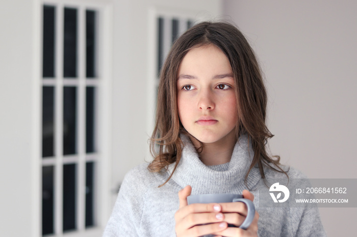 Close-up portrait of beautiful pensive tween girl in warm grey pullover holding cup of tea looking away. Natural beauty without make up.