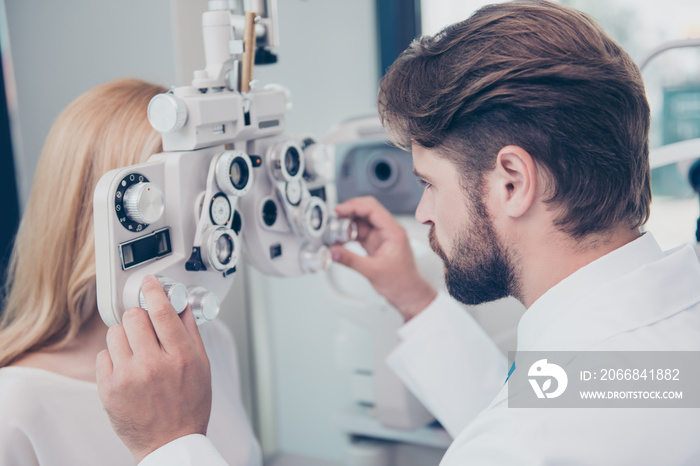 Close-up of brunet bearded doc optometrist adjusting phoropter for young blond female patient. Eye sight and healthcare