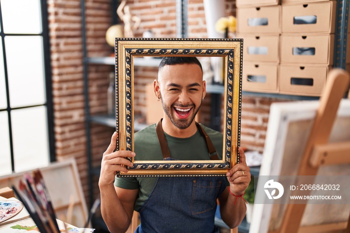 Young hispanic man sitting at art studio with empty frame smiling and laughing hard out loud because funny crazy joke.