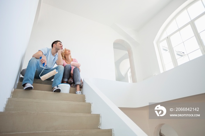 Low angle view of couple with painting tools sitting on steps in new house