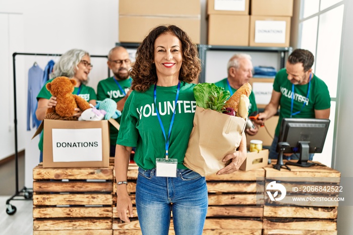 Group of middle age volunteers working at charity center. Woman smiling happy and holding paper bag with food to donate.