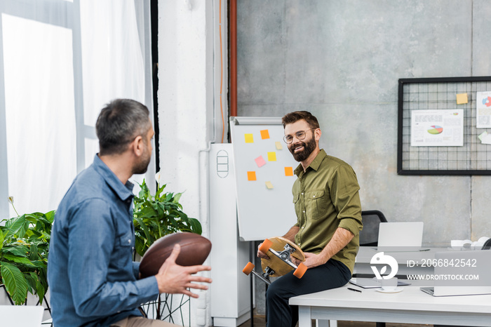 smiling businessmen looking at each other and holding american football ball and longboard in office