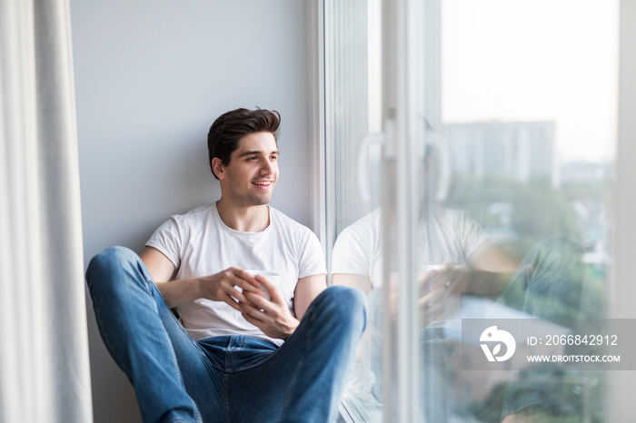 Handsome man sitting at window sill and drinking morning coffee