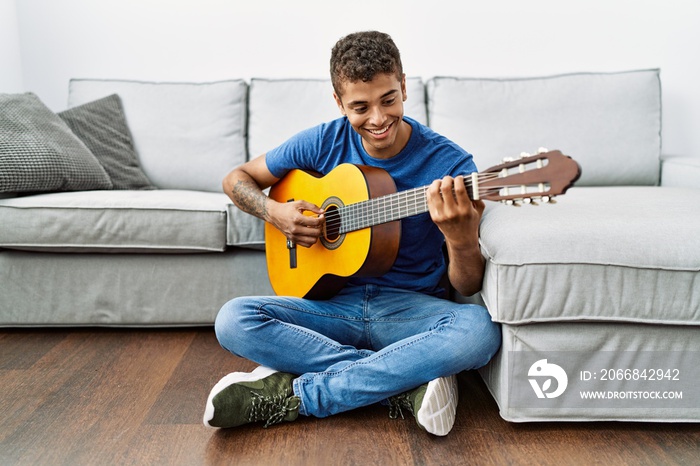 Young hispanic man playing guitar sitting on the floor at home