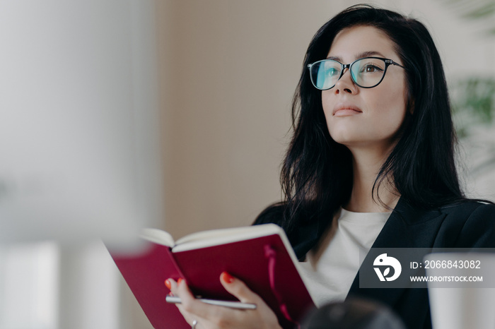 Beautiful woman with thoughtful expression, holds opened notepad, writes time management, makes plans for productive day, wears eyeglasses, poses indoor. Self employed female writes down ideas