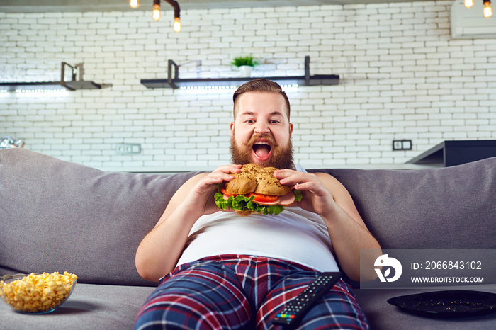 Fat funny man in pajamas eating a burger sitting on the sofa at home.