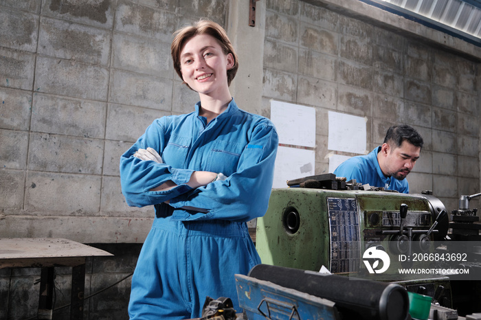 Portrait of Young White female industry worker stands, arms crossed, looks at camera and smiles, male partner works with machine behind her in manufacturing factory, professional mechanical engineers.