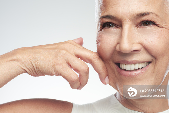 Gorgeous smiling Caucasian senior woman with short gray hair pinching her cheek. Beauty photography.