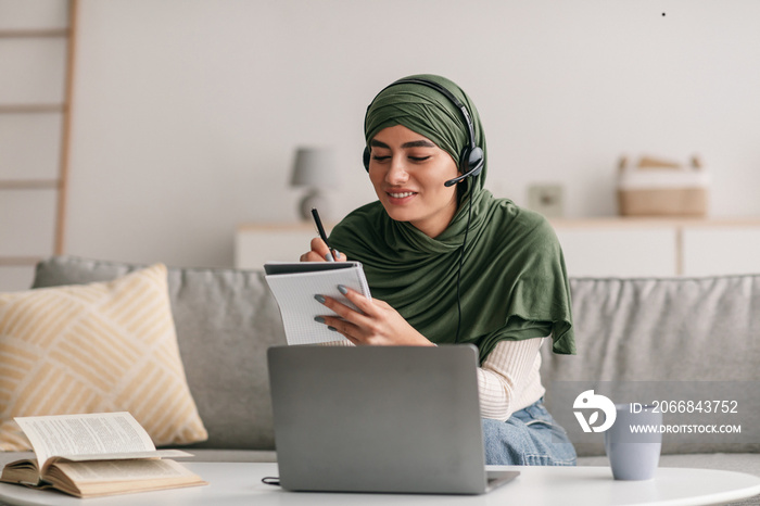 Cheerful Arab woman in hijab using laptop, wearing headphones, writing down information during remote lesson at home