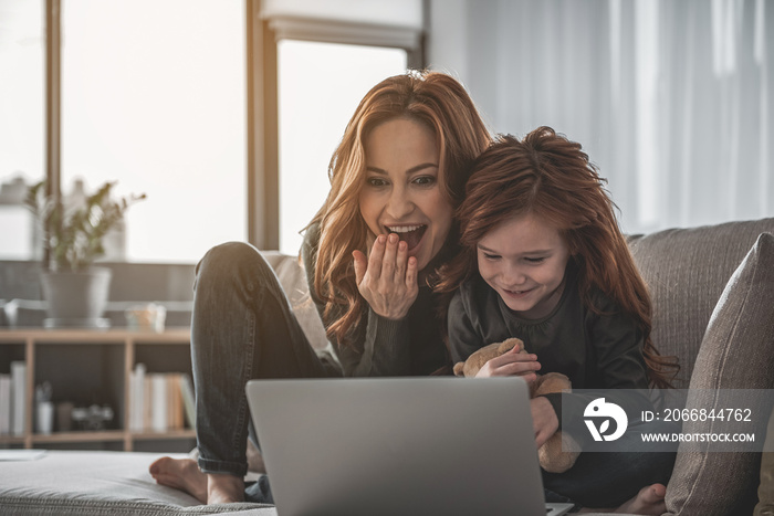 Excited woman is sitting on sofa with her kid holding teddy bear. They are watching something on computer and laughing with delight