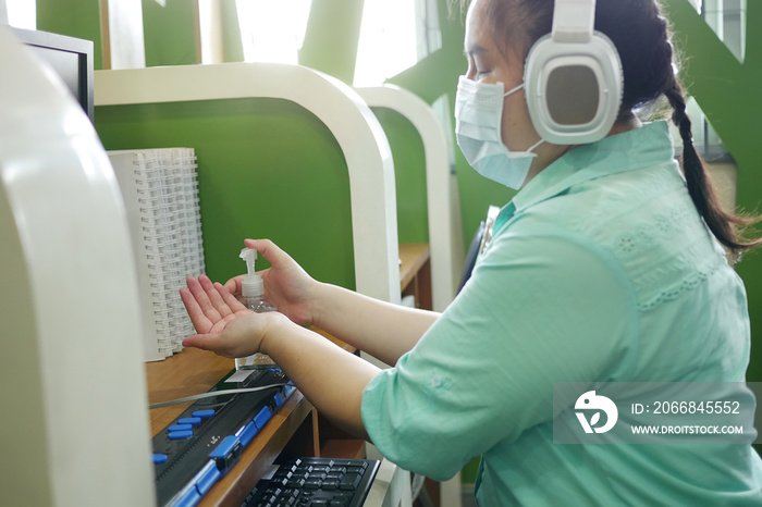 Disability blind person with headphone wearing face mask applying alcohol gel hand sanitizer on hands before using computer with braille display amid Coronavirus (COVID-19) pandemic.