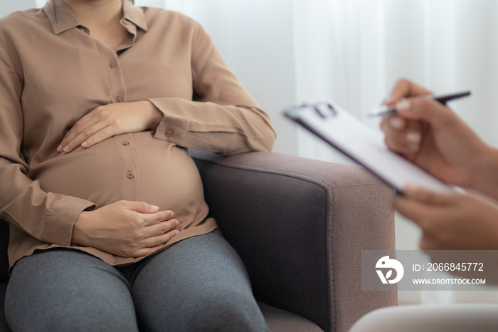 Asian young pregnant woman holding her belly while gynecologist notes the symptoms that the pregnancy is explaining about the unborn child.