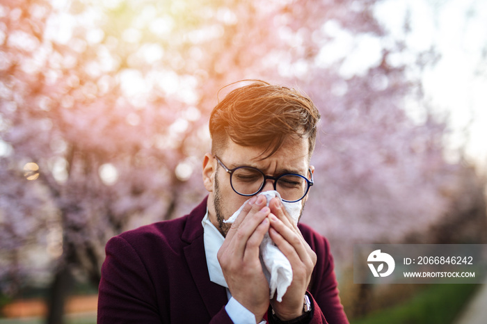 Young businessman sneezing in park. Allergy, flu, virus concept.