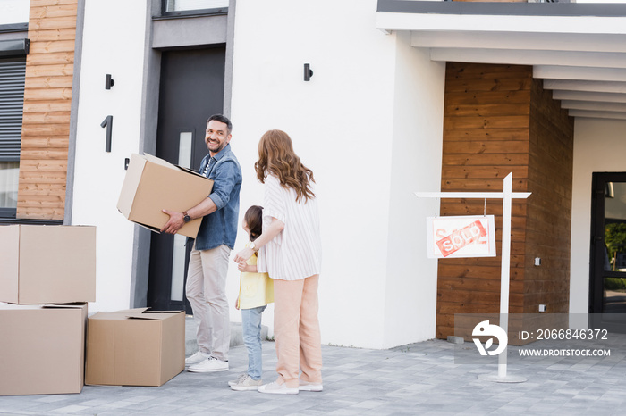 Smiling husband with cardboard box looking at wife holding hands with daughter near house and sign with sold lettering