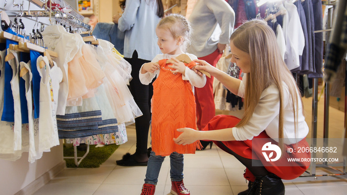 Cute little girl with mommy buying red dress in store of kids clothes