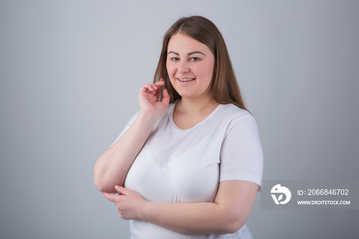 Beautiful plus size girl smiling in camera.Attractive white female model with long brown hair posing with toothy smile in studio wearing casual white t-shirt.Young cheeky woman with overweight smiles