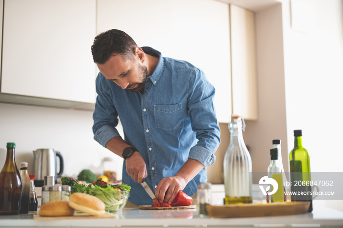 Waist up portrait of young gentleman in denim shirt cutting red bell pepper with knife while standing near table