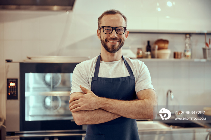 Cheerful bearded man in apron keeping arms crossed