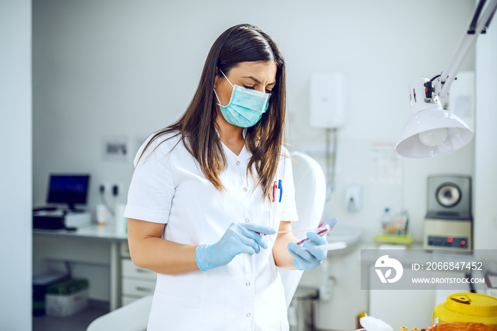 Dedicated caucasian lab assistant in white uniform, with protective mask and rubber gloves standing in lab and holding swab.