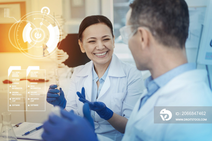 Positive working day. Cheerful happy scientist holding a test tube and smiling while looking at her colleague