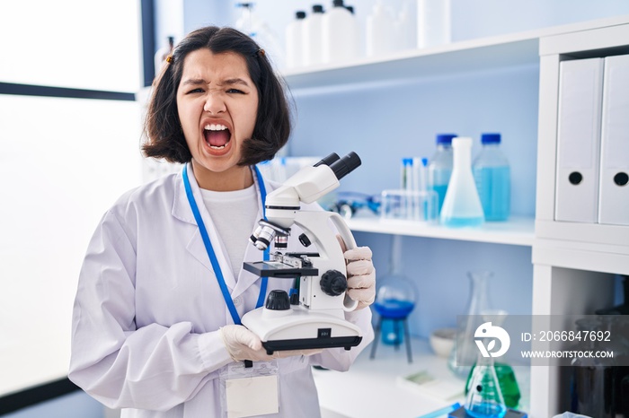 Young hispanic woman working at scientist laboratory holding microscope angry and mad screaming frustrated and furious, shouting with anger. rage and aggressive concept.