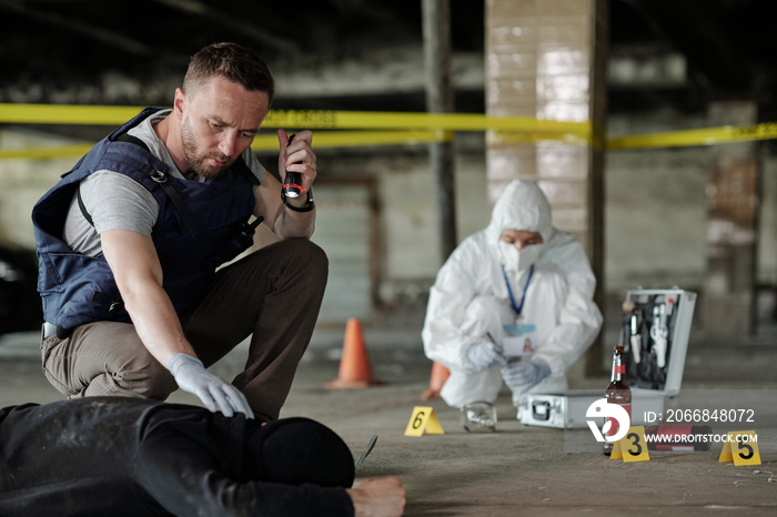 Detective inspector or policeman in bulletproof vest holding small lantern over dead body while inspecting crime scene in parking area