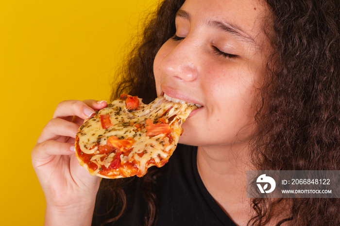 Brazilian, latin american girl for afro hair, yellow background, eating and biting delicious mini pizza, pizza, margarita pizza, cheese stretching, cheese.