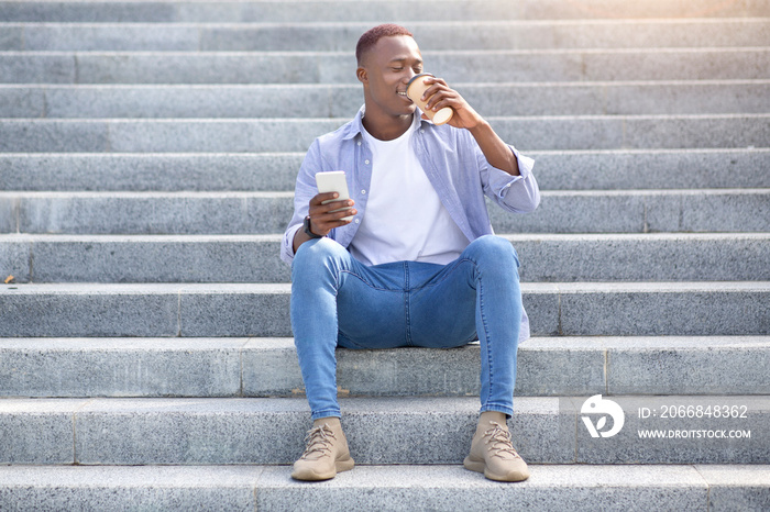 Handsome black man drinking takeaway coffee and using cellphone while sitting on stairs outside