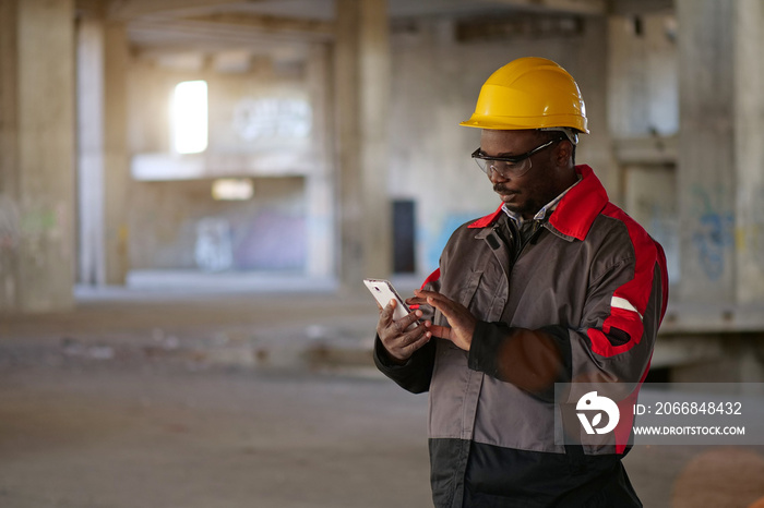 African american workman in yellow hard hat with smartphone