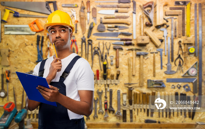 profession, construction and building concept- thinking indian worker or builder in helmet with clipboard and pencil over work tools on background