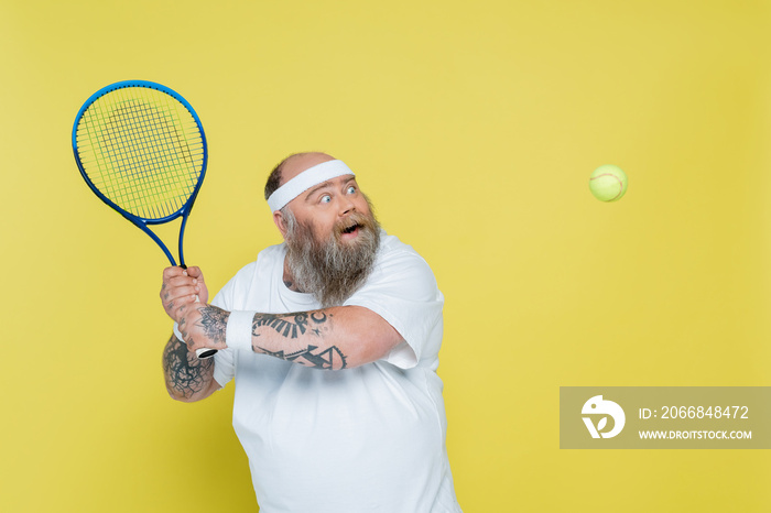 excited plus size man with tattoos playing tennis isolated on yellow.