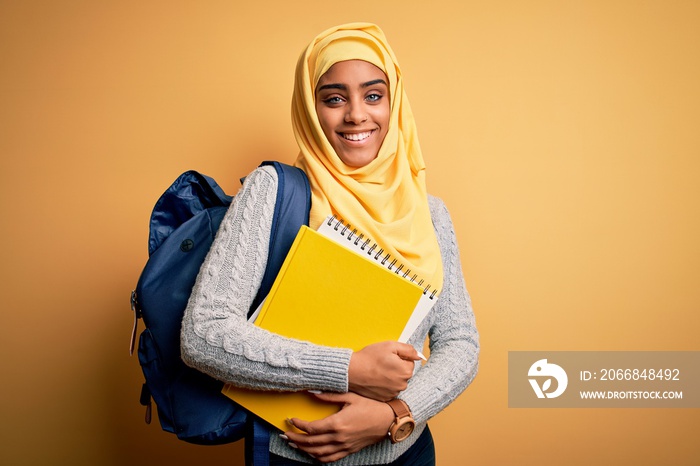 Young african american student girl wearing muslim hijab and backpack holding notebook with a happy face standing and smiling with a confident smile showing teeth