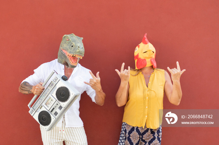 A crazy elderly couple wearing funny dinosaur and chicken masks. His holds a boombox. Background: red wall.