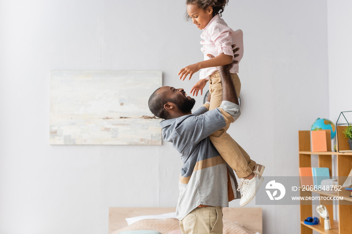 Side view of young African American man raising daughter above head while having fun at home