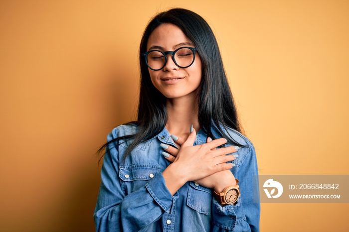 Young beautiful chinese woman wearing casual denim shirt over isolated yellow background smiling with hands on chest with closed eyes and grateful gesture on face. Health concept.