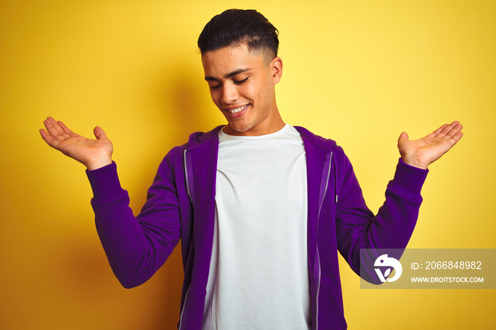 Young brazilian man wearing purple sweatshirt standing over isolated yellow background smiling showing both hands open palms, presenting and advertising comparison and balance