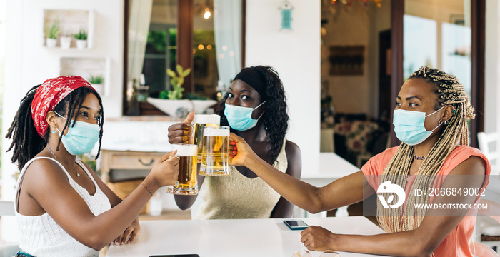 Three young African American girlfriends sitting at brewery (pub) with beer glasses wearing surgical masks. 3 Black girls are drinking ale in a luxury bar o restaurant. Toasting beer concept.