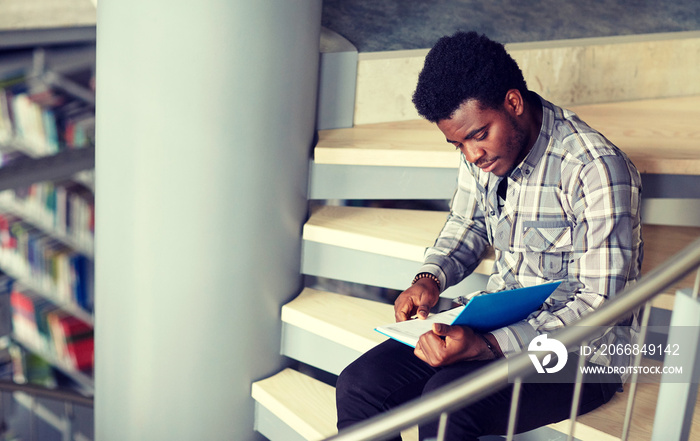 education, high school, university, learning and people concept - happy african american student boy or young man reading book sitting on stairs at library