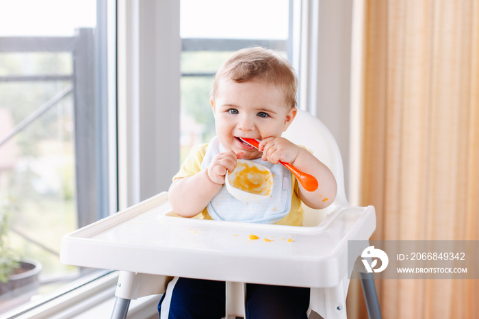 Portrait of cute adorable Caucasian child boy with dirty messy face sitting in high chair eating apple puree with spoon. Everyday home childhood lifestyle. Infant trying supplementary baby food