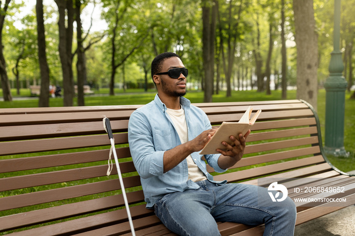 Young black visually impaired man sitting on bench in city park, reading Braille book outdoors