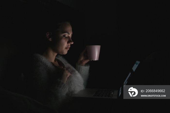 in the dark, the girl looks at the tablet pc screen and drinks coffee