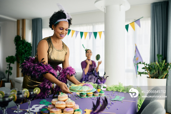 Happy black woman serving King cake at dining table during Mardi Gras celebration party at home.