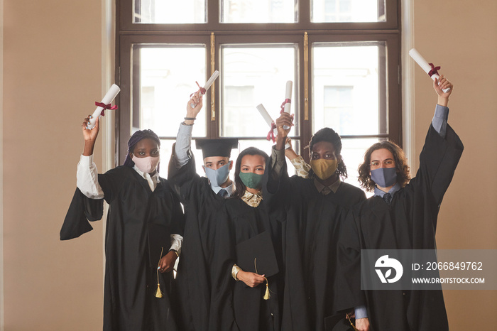 Diverse group of young people wearing masks and black ceremonial robes during indoor graduation ceremony in covid pandemic