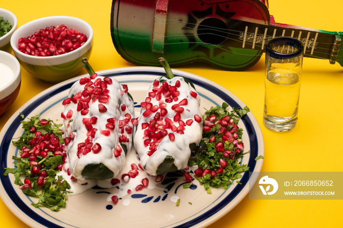 Chile en Nogada, a traditional dish from Puebla with the addition of walnut cream, pomegranate seeds and parsley, ingredients, toy guitar and glass of tequila on yellow background