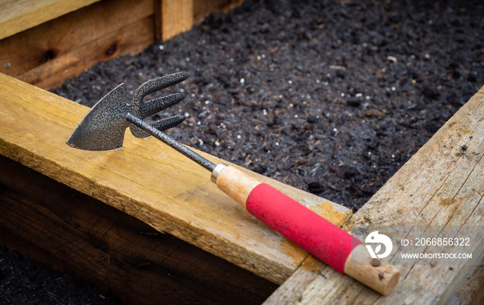 Wood and steel double sided hoe and rake garden hand tool on top of freshly planted wooden vegetable planter box.