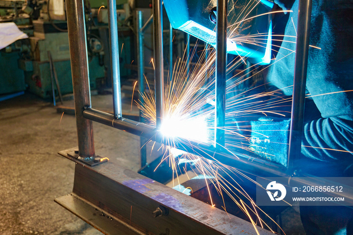 A welder at work in a workshop produces metal structures. Sparks from welding fly around.