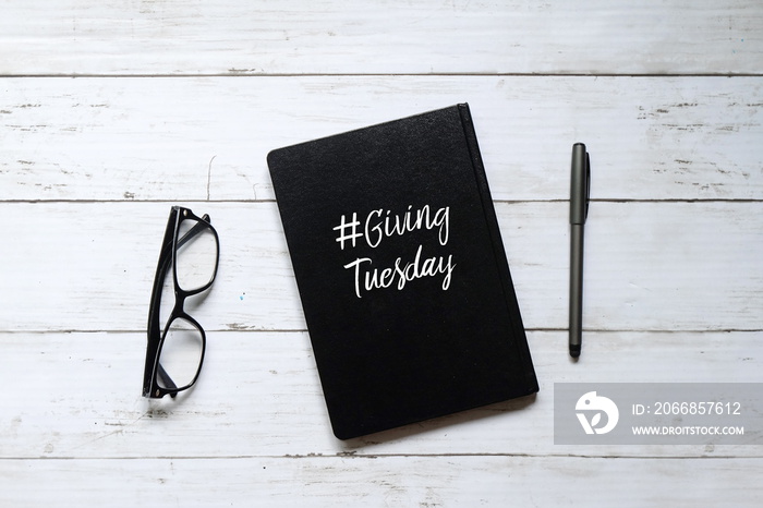 Top view of eyeglasses,pen and notebook written with ’#GIVINGTUESDAY’ on white wooden background.