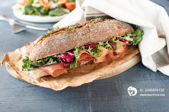 sandwich with baguette, ham, lettuce, kale on gray background, salad from kale and lettuce leaves and tomatoes on a white plate. healthy diet lunch, takeaway sandwich