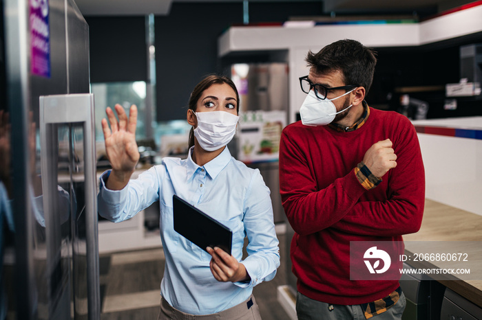 Young man talking with saleswoman about refrigerator he wants to buy. They are both with face protective masks. Pandemia, Covid-19 concept.