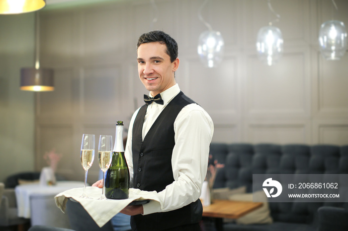 Young waiter holding tray with glasses and bottle of champagne indoors